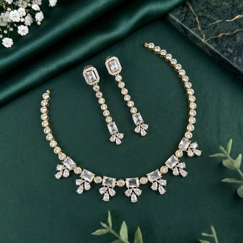 Gold-plated jewelry set featuring a necklace and matching drop earrings adorned with clear rectangular and round gemstones, arranged on a dark green satin fabric background with decorative floral and leaf props.