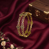 A pair of gold-plated Indian-style bangles decorated with small square pink gemstones and floral motifs, displayed on a draped white fabric with pink and white flowers in the background.