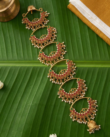 A gold-toned traditional Indian jhumka chain necklace with red and green stone embellishments and white bead drops, displayed on a green banana leaf.