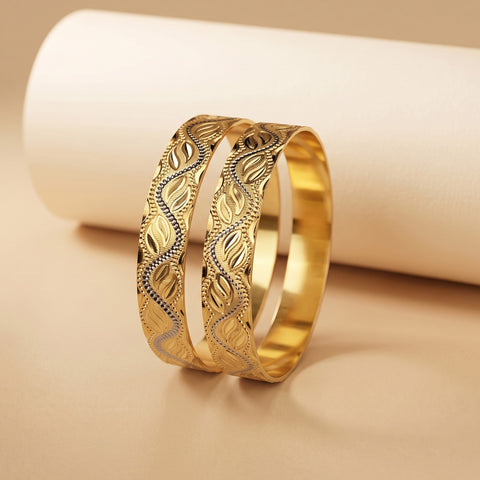 Two gold-toned bangles with intricate leaf and wave patterns, displayed on a beige surface with a white cylindrical prop.