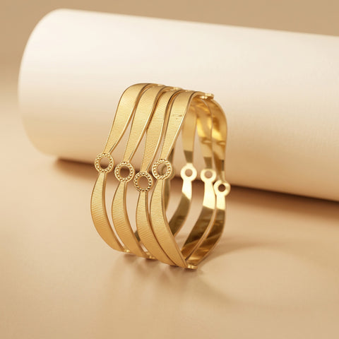 A stack of five gold-toned bangle bracelets with textured and smooth wavy bands, displayed on a beige surface in front of a white cylindrical prop.