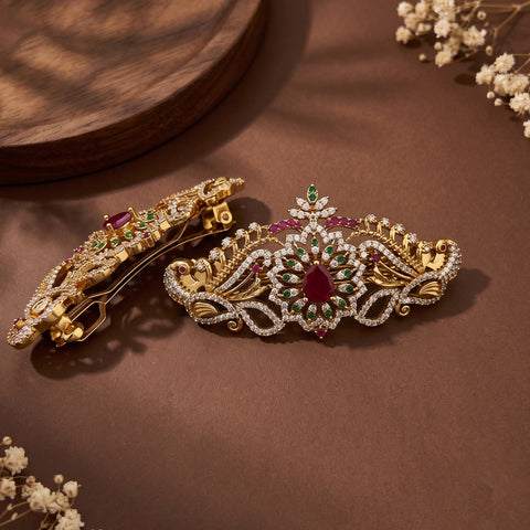 Two ornate gold hair clips decorated with red and green gemstones and clear rhinestones, displayed on a brown surface with decorative dried flowers.