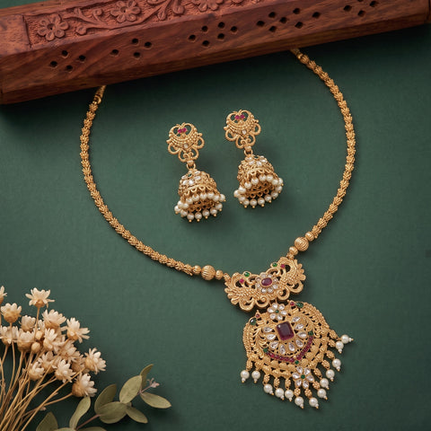 Gold-toned traditional Indian jewelry set featuring a necklace and matching earrings with pearl-like beads, red stone accents, and ornate filigree details, arranged on a dark green surface with a carved wooden prop and small dried flowers.