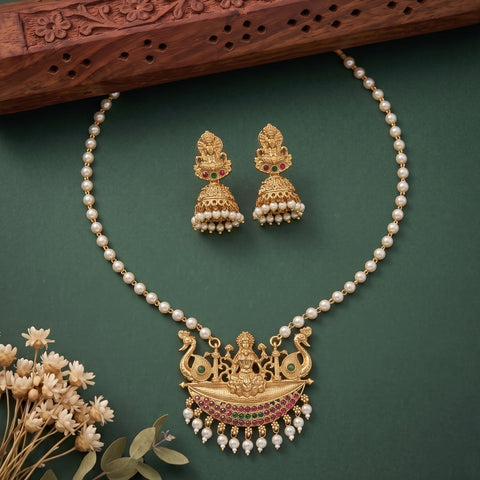 Gold and pearl jewelry set featuring a necklace and matching earrings with a detailed deity pendant, arranged on a dark green background with decorative dried flowers and a wooden prop.