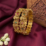 A set of three gold-plated floral bangles with small green and pink stone accents, displayed on draped burgundy fabric next to a carved wooden box and white flower buds.