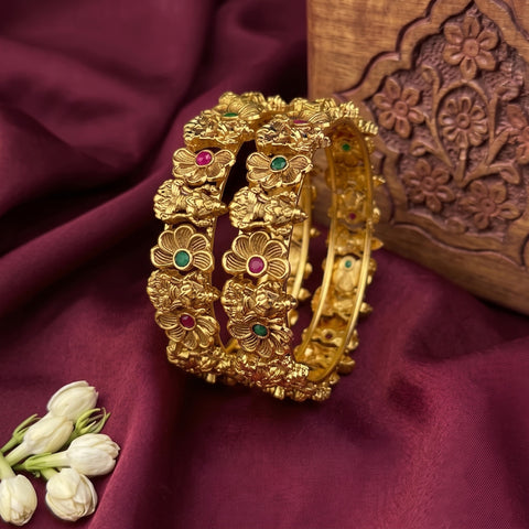 A set of three gold-plated floral bangles with small green and pink stone accents, displayed on draped burgundy fabric next to a carved wooden box and white flower buds.