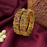 A pair of ornate gold bangles with green and pink stone accents, displayed on a deep burgundy satin fabric next to a carved wooden block.