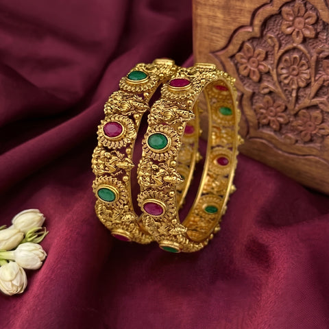 A pair of ornate gold bangles with green and pink stone accents, displayed on a deep burgundy satin fabric next to a carved wooden block.
