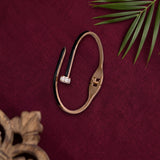 A gold-toned open cuff bracelet with a small clear gemstone accent, displayed on a textured dark red fabric background with decorative green leaves and a wooden object in the corners.