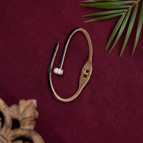 A gold-toned open cuff bracelet with a small clear gemstone accent, displayed on a textured dark red fabric background with decorative green leaves and a wooden object in the corners.