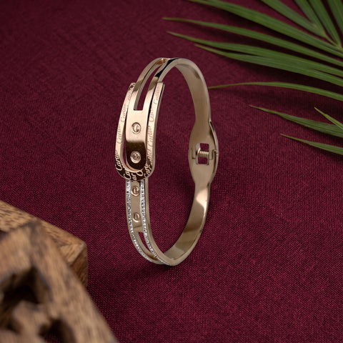 A rose gold bangle bracelet with a textured outer band, a polished inner band, and a row of small clear stones, displayed on a deep burgundy textured background with decorative leaves and a wooden block.