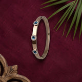 A slim metallic bangle bracelet with three circular blue evil eye motifs, displayed on a textured dark red fabric background with decorative palm leaves and a wooden object in the corners.