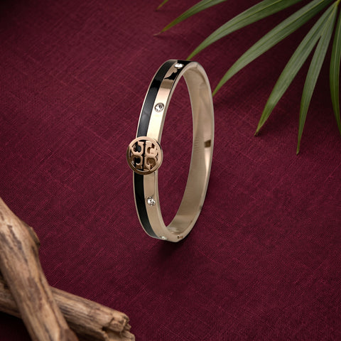 A slim bangle bracelet with a polished gold-tone finish, featuring a central round gold-tone medallion with an engraved monogram and small clear rhinestone accents, set against a deep burgundy textured background with decorative leaves and driftwood.