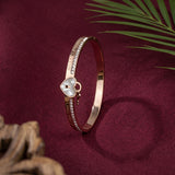A rose gold bangle bracelet with a heart-shaped mother-of-pearl charm and small clear crystals, displayed on a deep burgundy fabric background with decorative palm leaves and wood in the corners.