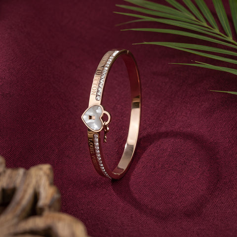 A rose gold bangle bracelet with a heart-shaped mother-of-pearl charm and small clear crystals, displayed on a deep burgundy fabric background with decorative palm leaves and wood in the corners.