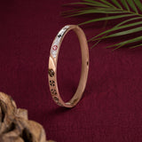 A slim rose gold bangle bracelet with white enamel inlays and small black and red symbols, displayed on a dark red textured fabric background with decorative leaves and wood in the corners.