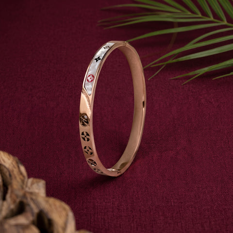 A slim rose gold bangle bracelet with white enamel inlays and small black and red symbols, displayed on a dark red textured fabric background with decorative leaves and wood in the corners.