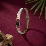 A metallic bangle bracelet with a black circular Chanel logo charm, set against a deep burgundy fabric background with decorative green leaves and a wooden object.