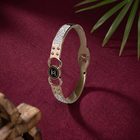 A metallic bangle bracelet with a black circular Chanel logo charm, set against a deep burgundy fabric background with decorative green leaves and a wooden object.