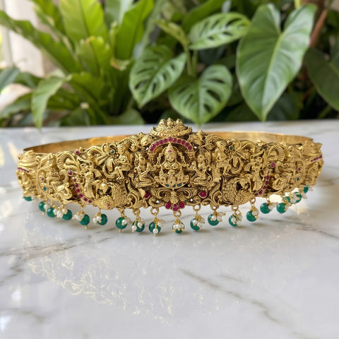 An ornate gold-toned traditional Indian headpiece (tilak) decorated with intricate carvings, red gemstone accents, and dangling green and white bead tassels, displayed on a white marble surface with a blurred green plant background.