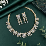 Gold-toned jewelry set with a matching necklace and earrings, featuring clear square and teardrop stones set in ornate geometric and floral motifs, arranged on dark green satin fabric with small decorative flowers and leaves.