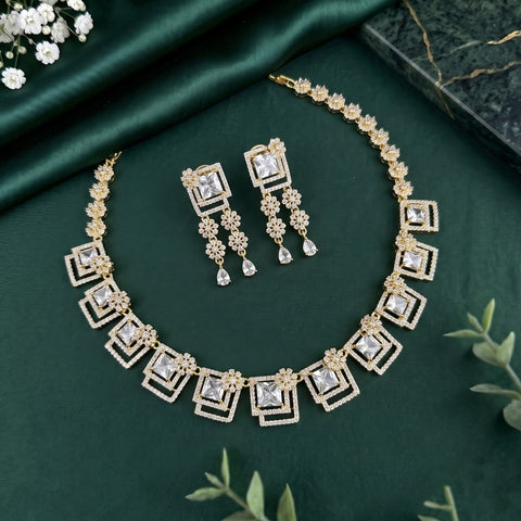Gold-toned jewelry set with a matching necklace and earrings, featuring clear square and teardrop stones set in ornate geometric and floral motifs, arranged on dark green satin fabric with small decorative flowers and leaves.