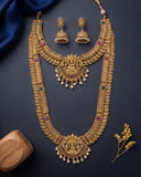 An ornate gold-toned traditional necklace set with matching earrings, decorated with small multicolored stones and white pearl-like beads, arranged on a dark navy background with decorative fabric and a wooden block.
