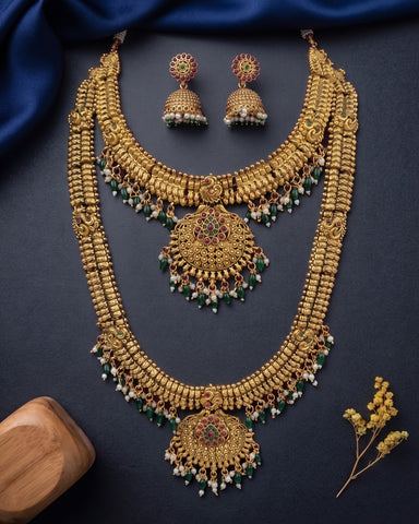 An ornate gold-toned traditional Indian jewelry set featuring a double-strand necklace with a large pendant, matching earrings, and small dangling bead accents in green, white, and red, arranged on a dark navy fabric background with decorative props.