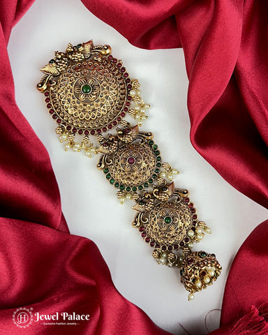 Gold jewelry piece with green and red stones on a red fabric background, brand 'Jewel Palace' visible.