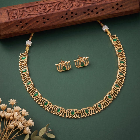 Gold-toned necklace and matching earrings set with green stone accents, arranged on a dark green background with a carved wooden prop and small dried flowers.