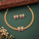 Gold-tone butterfly jewelry set featuring a choker necklace and matching stud earrings, decorated with pink and green stones and white crystal-like accents, displayed on a dark green background with decorative dried flowers and a carved wooden block.