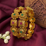 A pair of gold-plated traditional Indian bangles decorated with floral and square motifs, accented with green stone and pink stone details, displayed on a draped white fabric background.