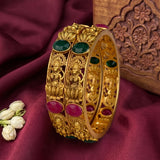 An ornate gold-plated triple bangle bracelet decorated with green and pink gemstone accents and detailed deity carvings, displayed on a draped maroon fabric next to white jasmine buds.