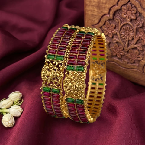 Three gold-plated Indian bangles with rectangular red stones and green stone accents, displayed on a draped burgundy fabric with a carved wooden block and white flowers as props.