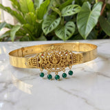 An ornate gold-toned traditional Indian headpiece (tilak) with intricate filigree work, a central deity motif, and four dangling green bead drops, displayed on a white marble surface with a blurred background of green plants.