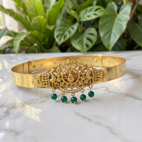 An ornate gold-toned traditional Indian headpiece (tilak) with intricate filigree work, a central deity motif, and four dangling green bead drops, displayed on a white marble surface with a blurred background of green plants.