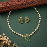 Gold-plated necklace and matching earrings set with white pearls and green stone paisley motifs, displayed on a dark green background with dried flowers and a carved wooden prop.