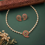 Gold-plated necklace and matching earrings set with white pearl beads and red and green paisley-shaped stone accents, displayed on a dark green background with a carved wooden prop and small dried flowers.