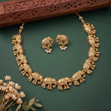 Gold elephant necklace and matching stud earrings displayed on a dark green background, with small dried flowers at the lower left and a carved wooden prop at the top.
