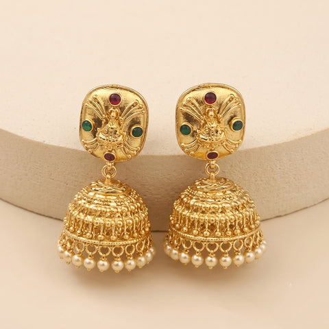 A pair of gold-toned Indian jhumka earrings with detailed filigree bells, small white pearl beads along the bottom, and small red and green stone accents on the studs, displayed on a beige curved stand.