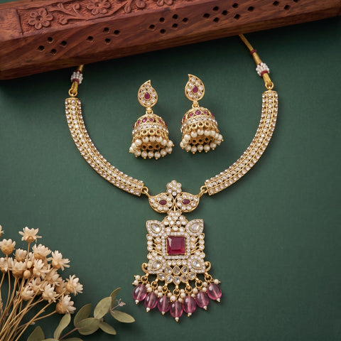 Gold-plated Indian traditional jewelry set featuring a necklace and matching earrings with white faux pearls, red gemstone accents, and pink crystal drops, displayed on a dark green background with decorative dried flowers and a wooden block.