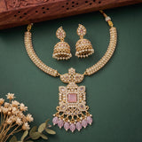 Gold-plated Indian bridal jewelry set featuring a necklace and matching earrings with white faux pearls and pink gemstone accents, displayed on a dark green background with decorative dried flowers and a wooden block.