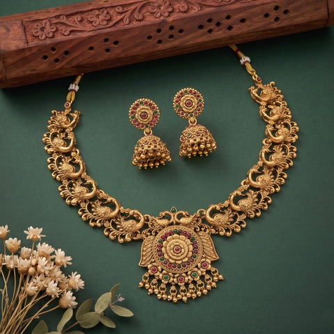 An ornate gold-toned traditional necklace with matching earrings, displayed on a dark green background with a carved wooden prop and small dried flowers. The jewelry features detailed filigree, peacock motifs, and floral elements accented with red, green, and pink stones.