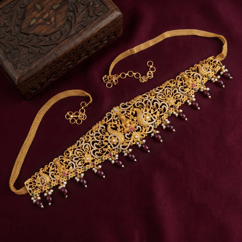 An ornate gold-toned traditional necklace with intricate filigree detailing, small red gemstone accents, and dangling pearl-like beads, displayed on a deep burgundy fabric background next to a carved wooden box.