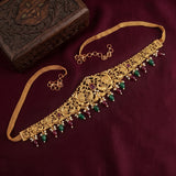 An ornate gold-toned traditional necklace choker with intricate filigree detailing, decorated with green and purple teardrop beads and small white pearls, displayed on a dark burgundy fabric next to a carved wooden box.