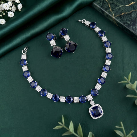 A jewelry set featuring a necklace and matching earrings with deep blue gemstones and silver-toned settings, arranged on a dark green satin fabric background with decorative greenery and flowers.