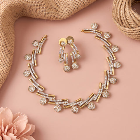 Gold and crystal jewelry set featuring a curved necklace, matching drop earrings, and a small gold stud, arranged on a soft pink background with burlap and a flower prop.