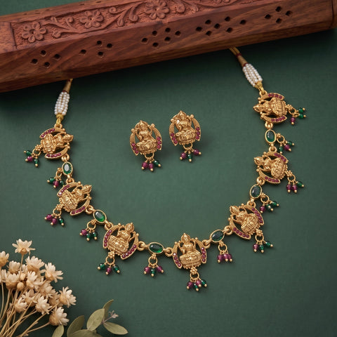 Gold-toned jewelry set featuring a necklace and matching earrings with detailed deity motifs, red and green stone accents, and pearl bead details, arranged on a dark green background with decorative dried flowers and a carved wooden prop.