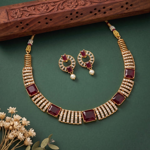 Gold-plated Indian style jewelry set featuring a choker necklace and matching earrings with square dark red gemstones and rows of clear stones, displayed on a dark green background with a carved wooden prop and dried flowers.