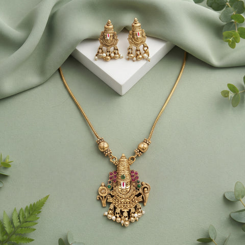 Gold-toned traditional necklace and matching earrings displayed on a sage green fabric background with decorative green leaves and a white display block.